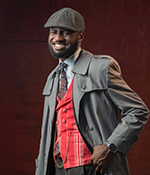 male with black hair in gray top coat and cap, red vest
